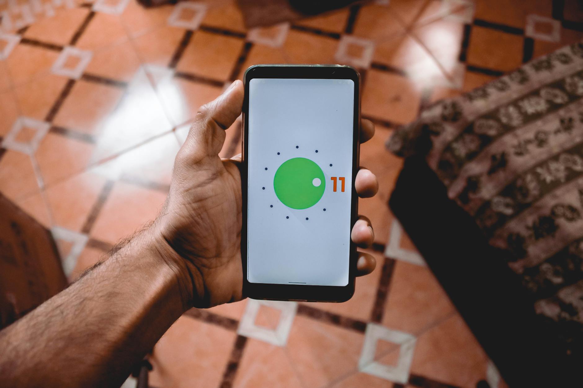 Dłoń trzymająca smartfon z interfejsem Android 11 nad wzorzystą podłogą
